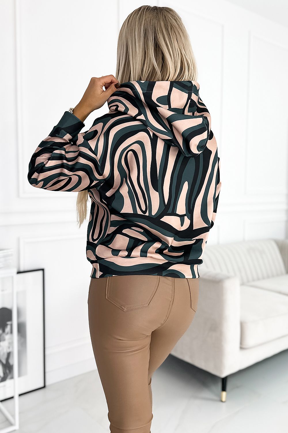 Vrouw in een hoodie met abstract zwart-groen-beige patroon, gecombineerd met beige leren broek.achter uit zicht