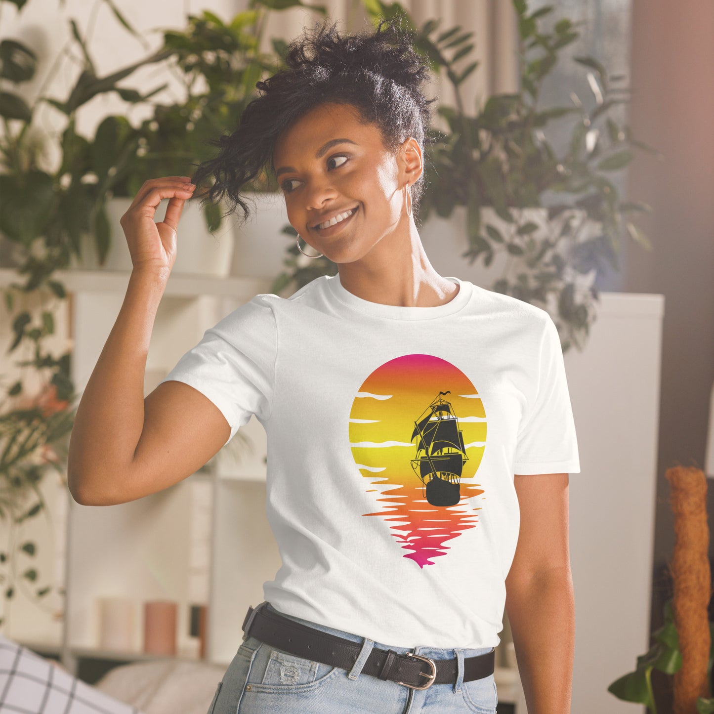 Een unisex basic softstyle T-shirt met een afbeelding van een zonsondergang en een zeilboot, waar een vrouw naar links kijkt, met een huiselijke sfeer op de achtergrond.