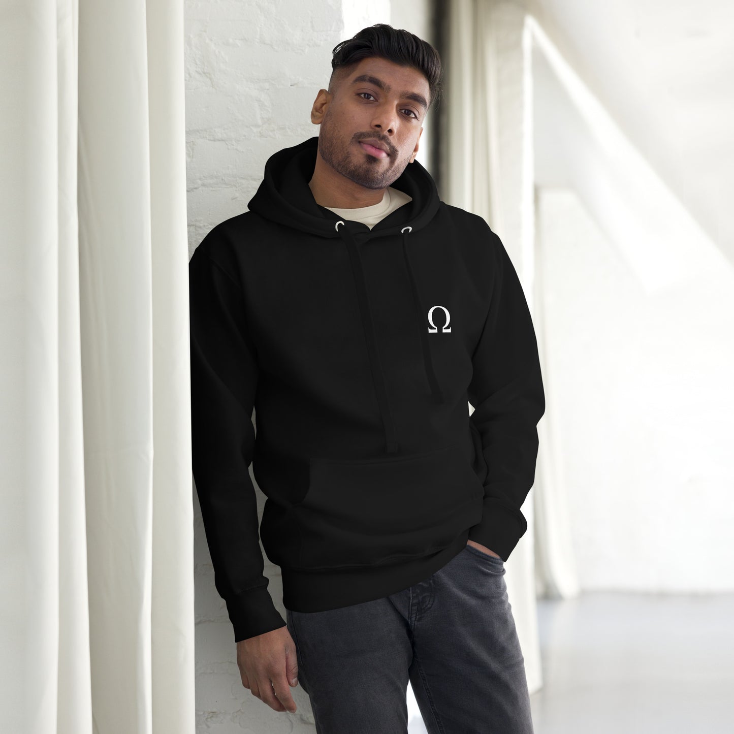 Een zwarte unisex premium hoodie met het Omega-symbool op de voorkant, gedragen door een man tegen een witte achtergrond, met een subtiel huisstijlaccent van gordijnen