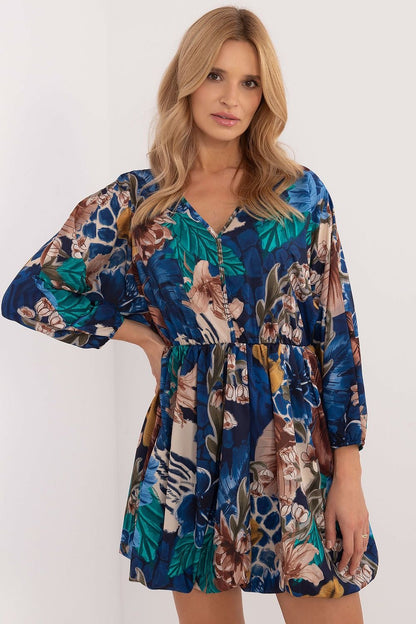 Een vrouw draagt een korte, losvallende jurk met lange mouwen en een opvallende print van kleurrijke bloemen en subtiele dierenmotieven in blauwtinten,voor uitzicht