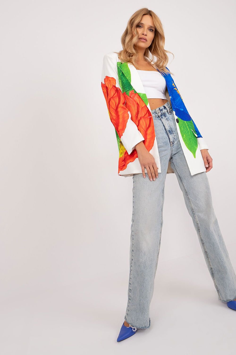 Vrouw draagt een witte blazer met grote, kleurrijke bloemenprint, gecombineerd met een witte crop top en jeans. Witte achtergrond