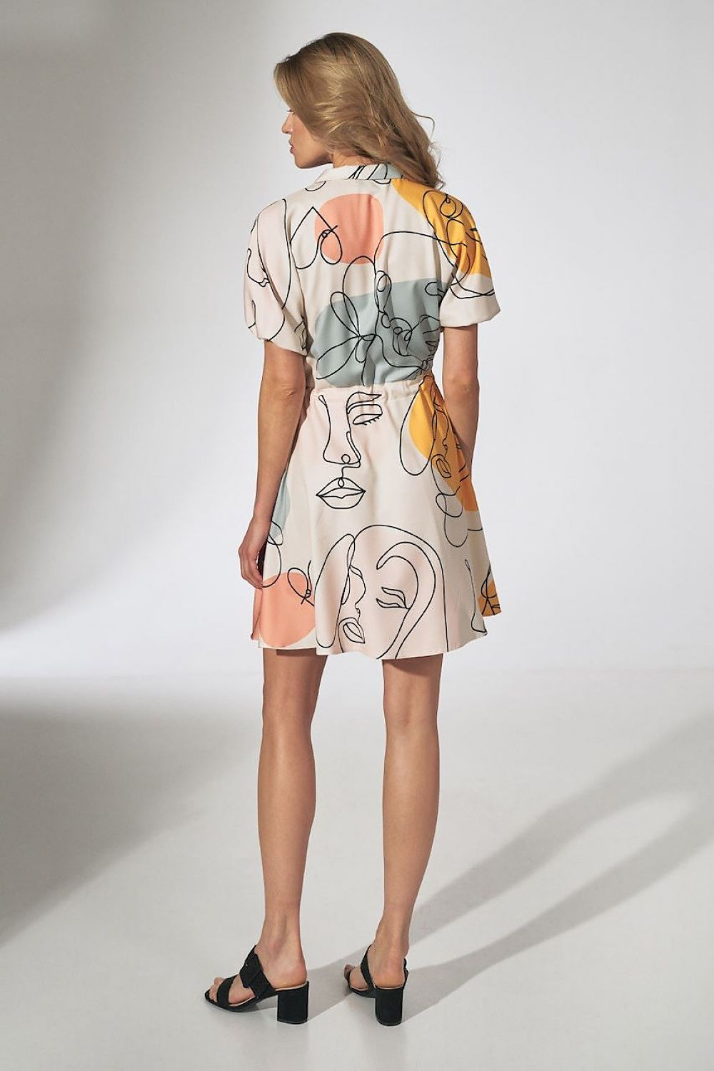 Vrouw gezien van achteren draagt een korte, luchtige zomerjurk met een abstracte gezichtenprint in pastelkleuren, korte mouwen en een strikceintuur, gecombineerd met zwarte sandalen met hak.acheraanzicht